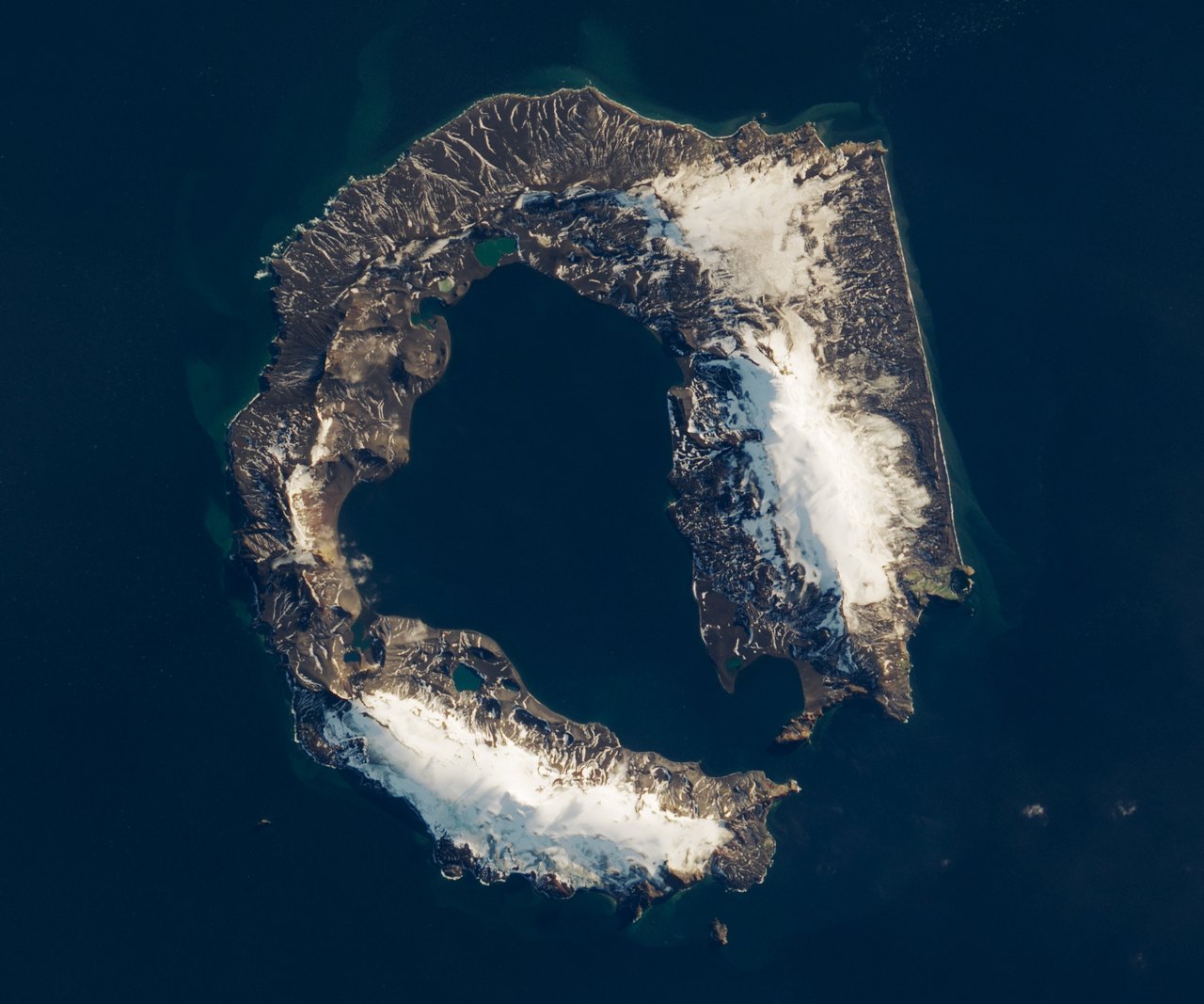 Natural-color Landsat view of horseshoe-shaped Deception Island, where dark volcanic slopes and snow surround a flooded caldera.
