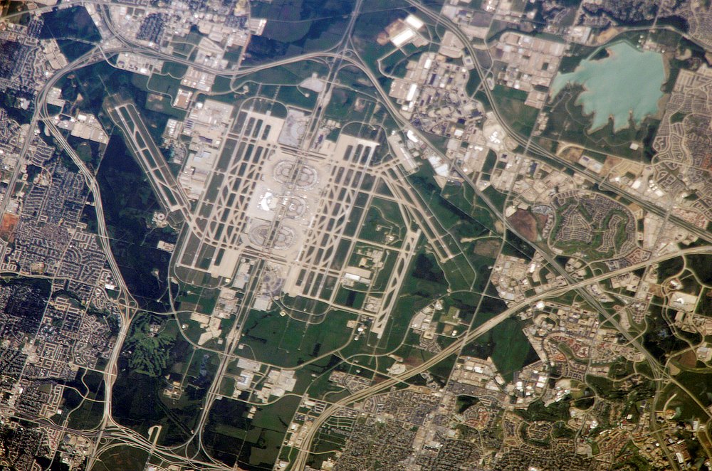 Oblique astronaut view of Dallas–Fort Worth airport, with parallel runways and terminal buildings amid roads, fields, and neighborhoods.