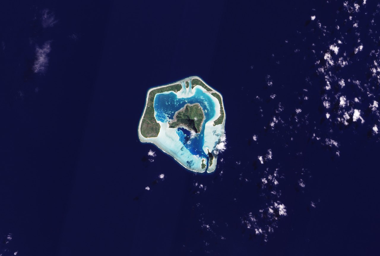 True-color satellite view of Maupiti, with a mountainous central island, blue lagoon, pale encircling reef, and surrounding dark Pacific.
